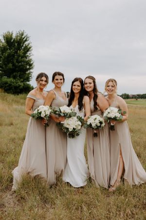 Bridal party wearing neutral beige gowns and holding white floral bouquets pose together in a grassy field.