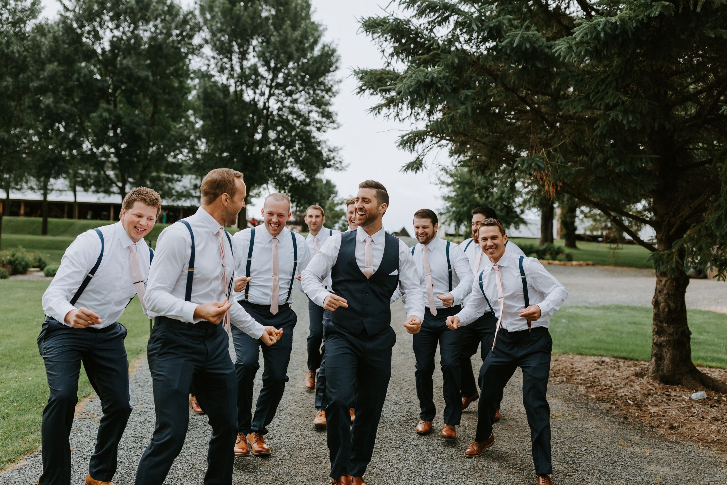 Groomsmen in suspenders and dress shirts pose playfully outdoors.