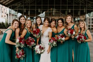 Bridesmaids in elegant emerald green dresses hold burgundy and white bouquets while posing with bride in greenhouse.