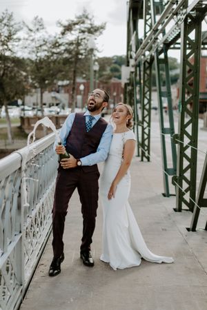 Wedding couple laughing together on a bridge with industrial architecture in the background.