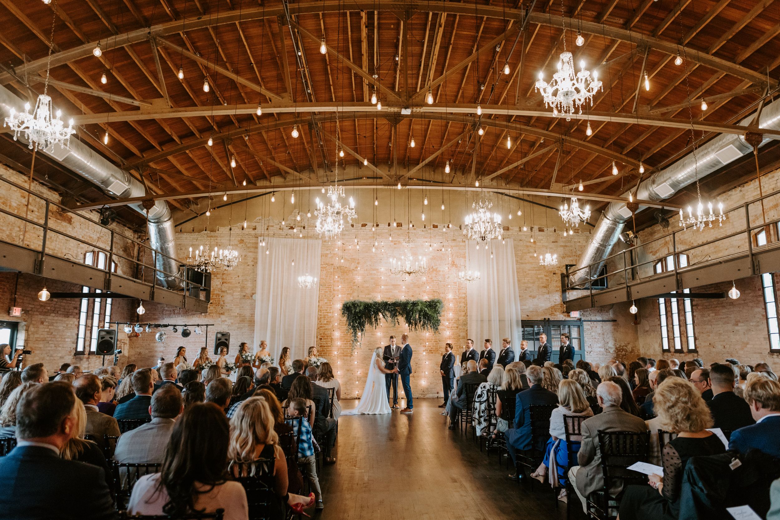 An indoor wedding ceremony in a rustic venue with exposed beams and hanging lights.