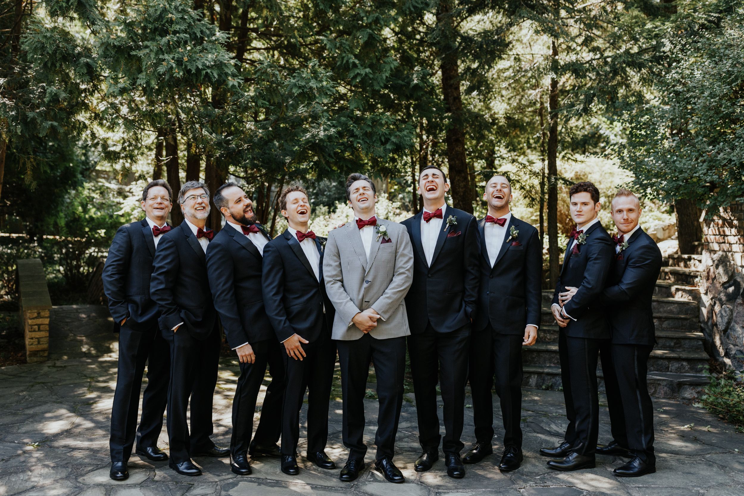 A group of groomsmen in matching dark suits pose outdoors at a wedding.