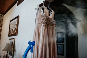 Blush bridesmaid dresses hang elegantly on wooden hangers against a white brick wall.