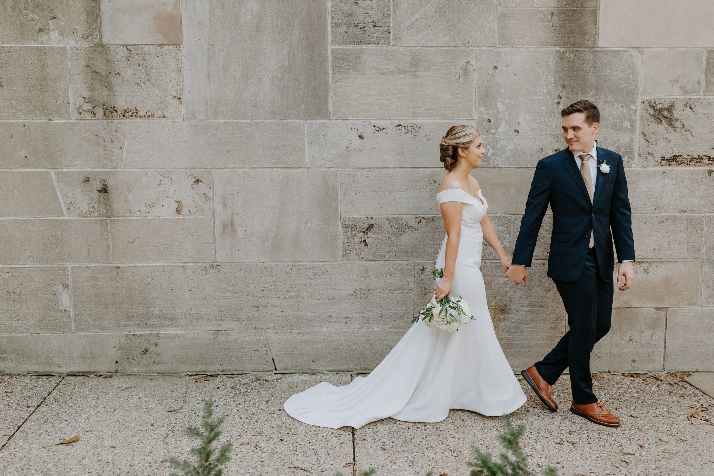 Elegant couple poses against stone wall background in wedding attire.