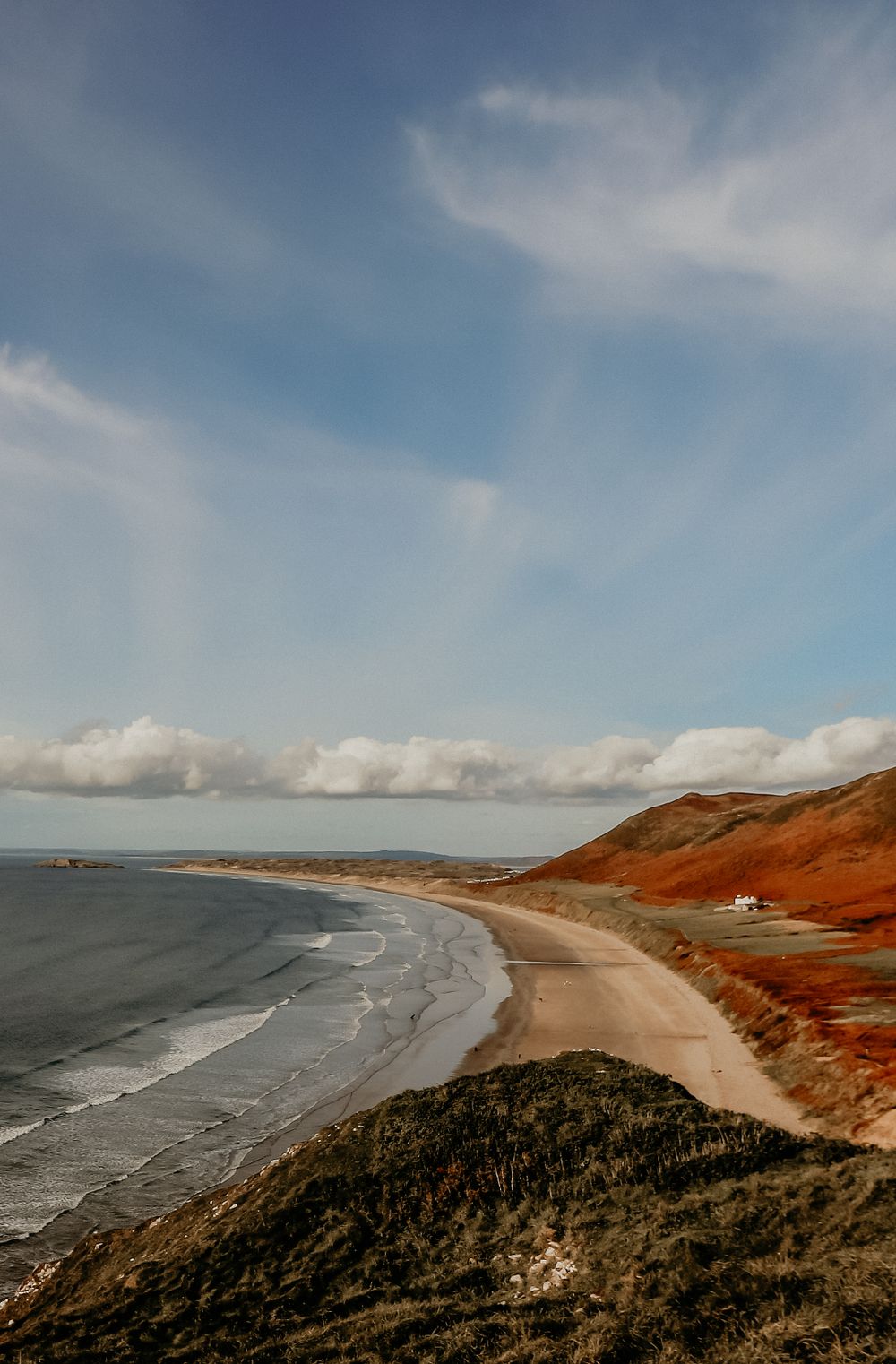 Dramatic coastline view with curved beach, red cliffs, and wispy clouds in the blue sky.