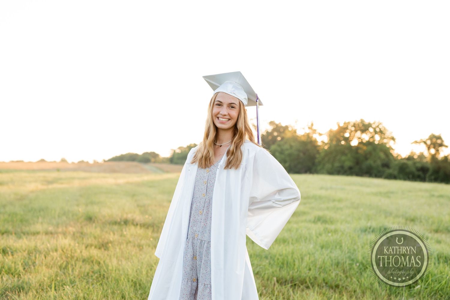 Graduate in white gown poses in sunny field during golden hour.
