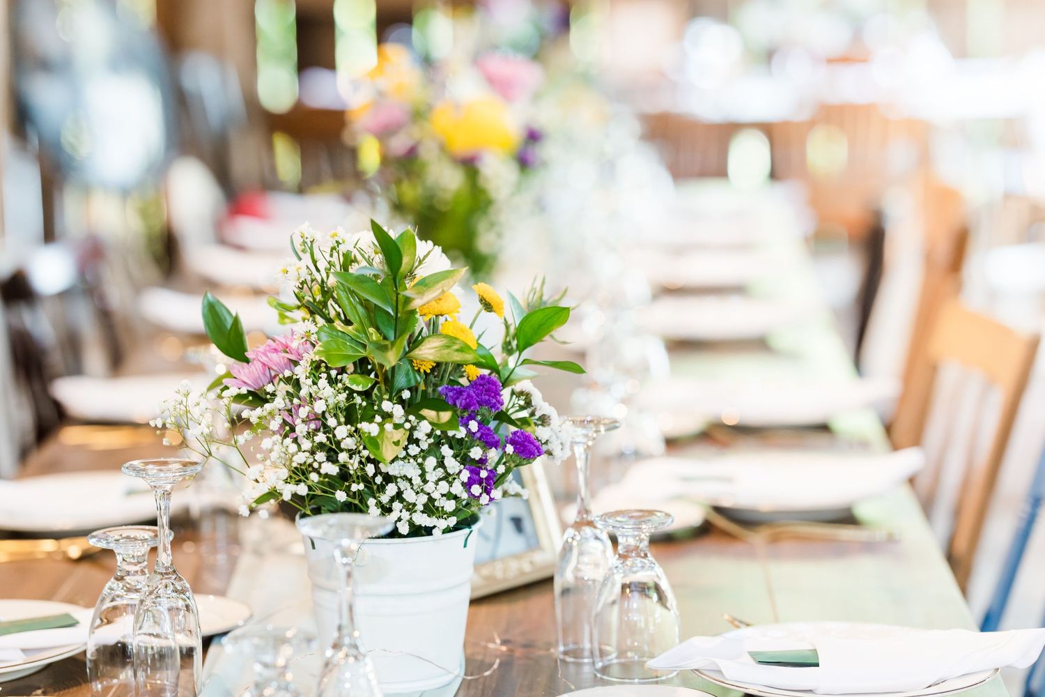 A rustic wedding table setting with wildflower centerpieces in white buckets and elegant glassware.