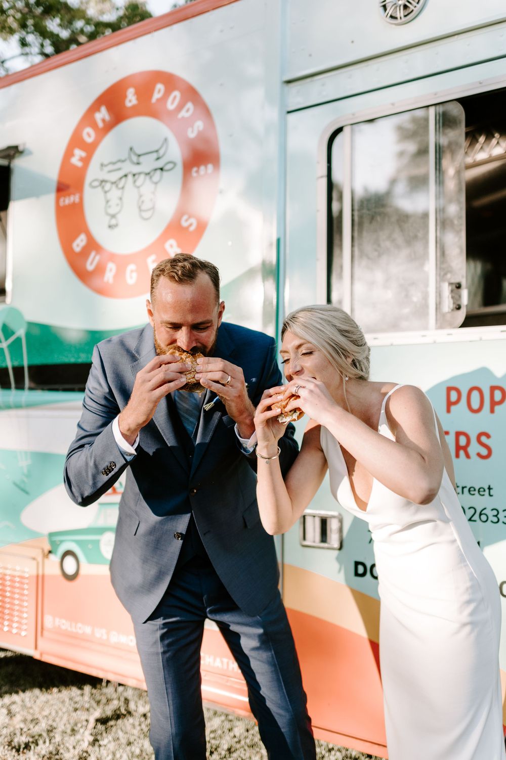 A couple takes big bites of food while standing in front of a mint and coral food truck.