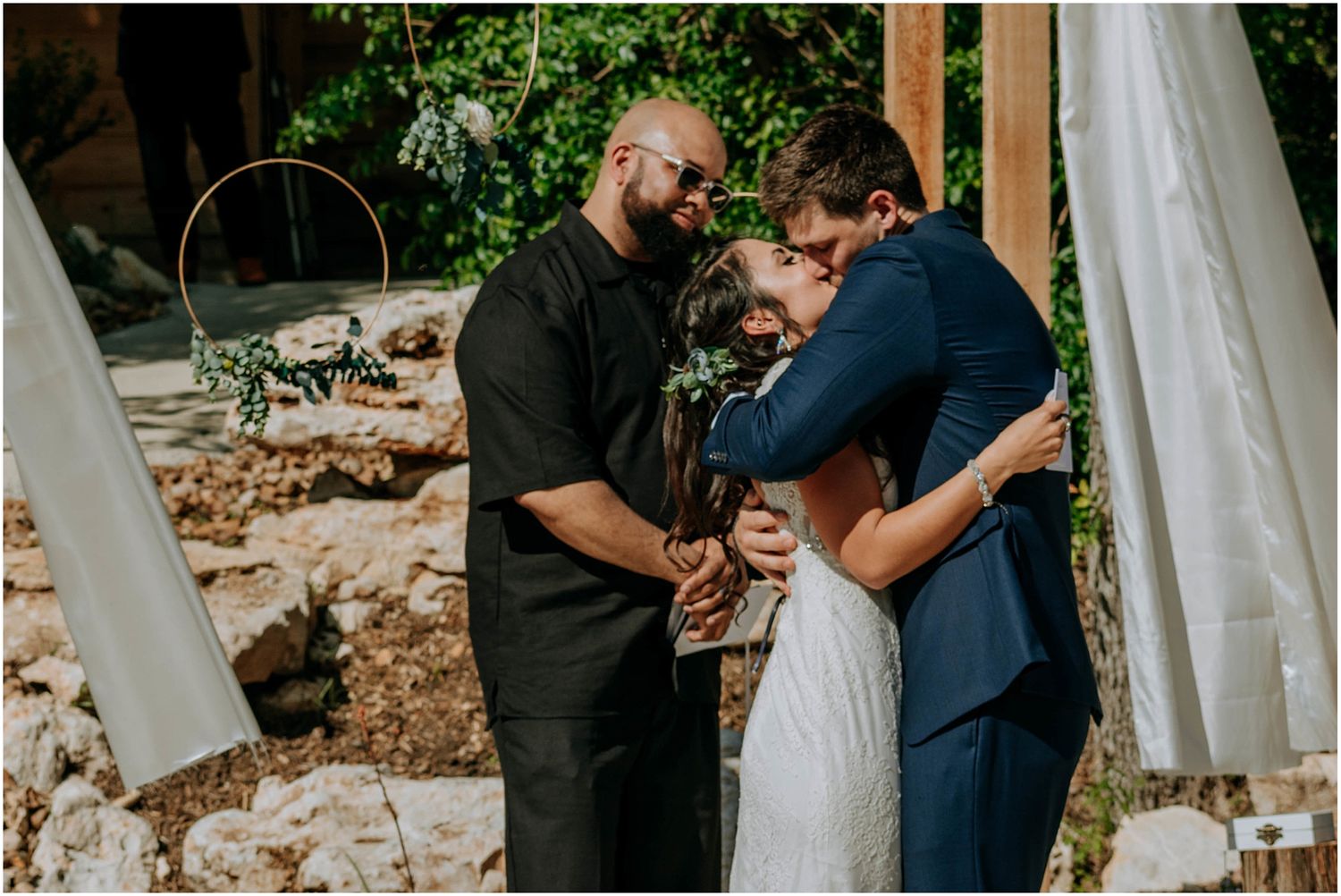 A tender moment during an outdoor wedding ceremony beneath a decorated wooden arch.