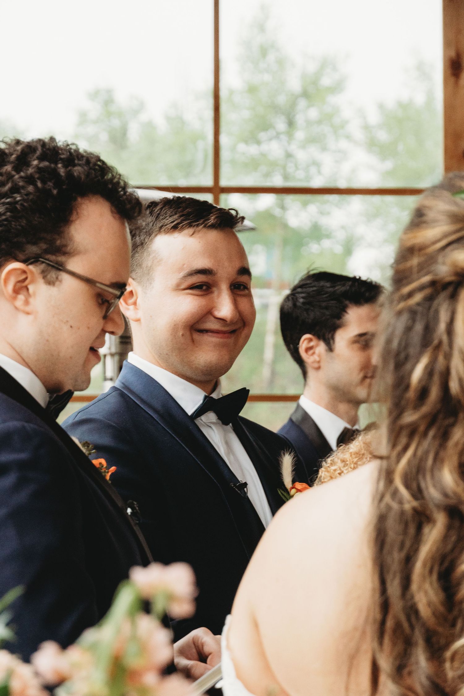 Wedding ceremony with groomsmen in formal attire standing by large windows overlooking nature.