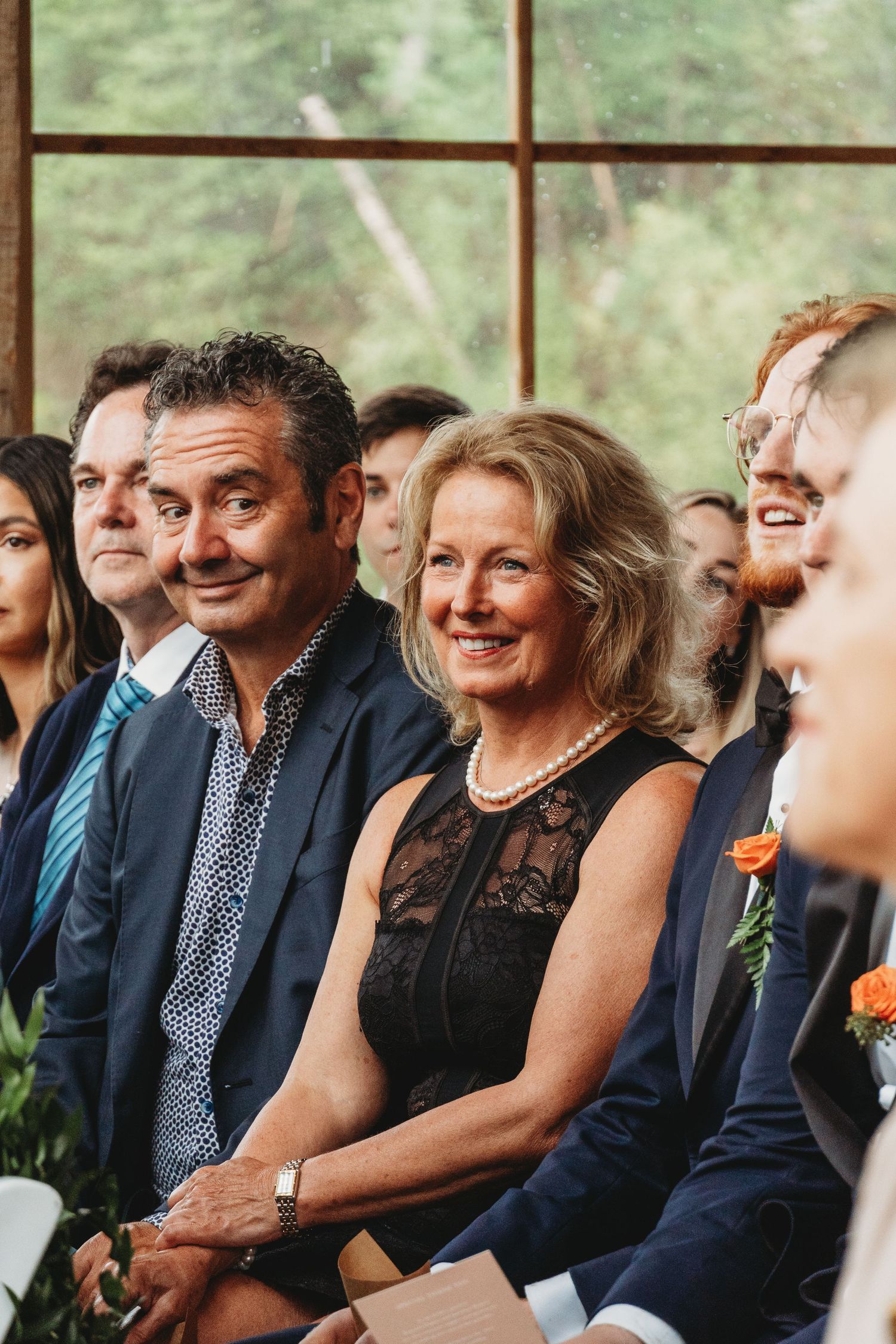 Wedding guests smiling during ceremony in front of windows with natural forest backdrop.
