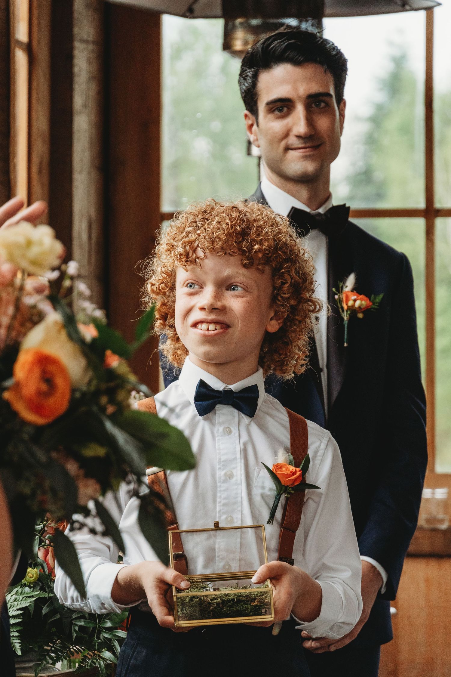 Young ring bearer in bow tie and suspenders stands in front of formal wedding backdrop with floral arrangements.