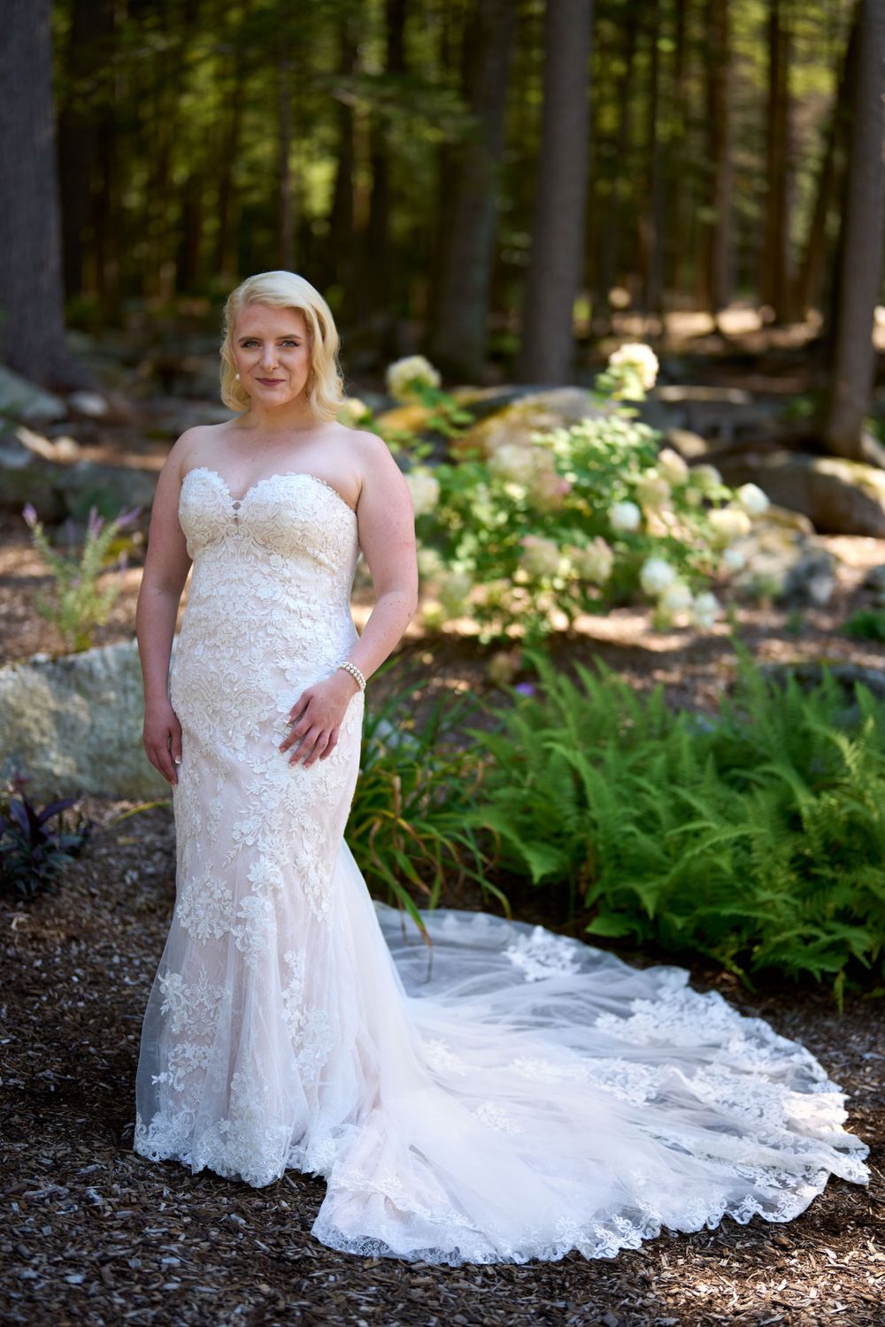 bride in strapless wedding gown and full lace train stands in green ferned woodland
