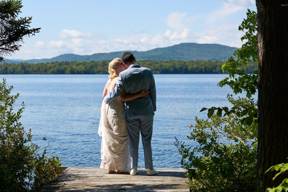 Bride in lacy white gown embraces her blue suited groom by shore of Squam Lake