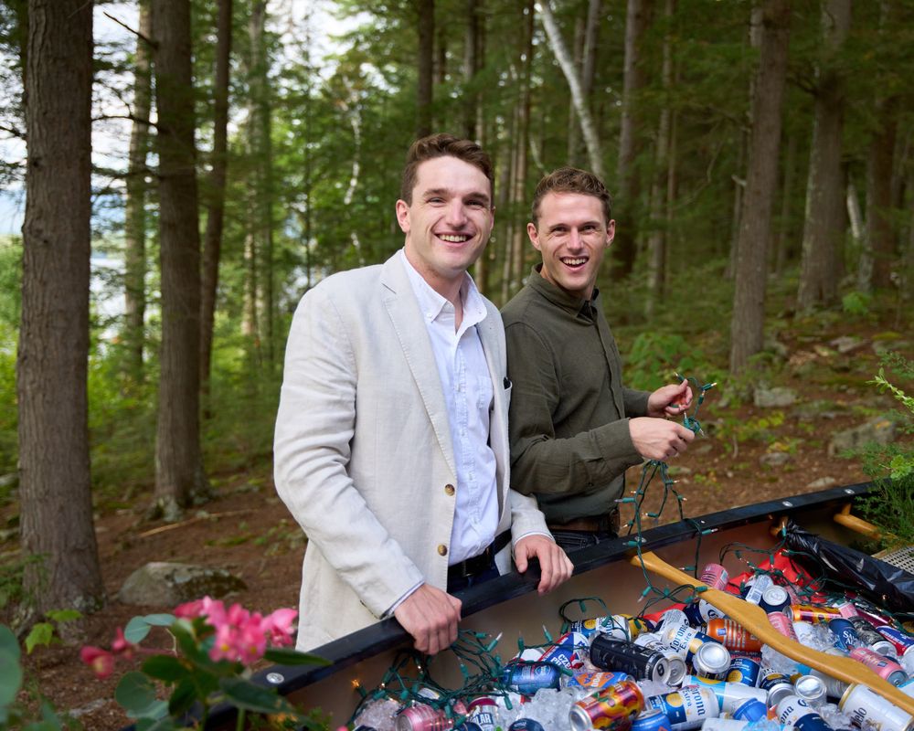 Two young men stand by the side of a canoe full of wedding reception drinks