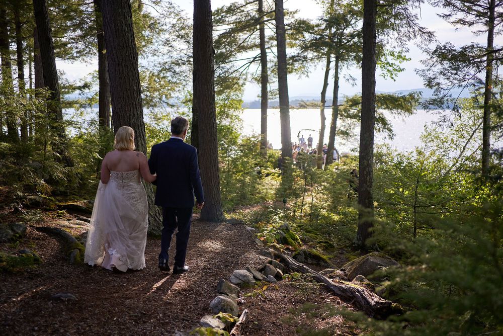 Amidst tall evergreen trees by Squam Lake shore, bride and groom stroll pine needled path