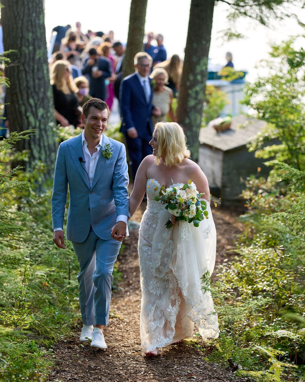 Smiling newlyweds on woodland path with wedding guests behind them Squam Lake