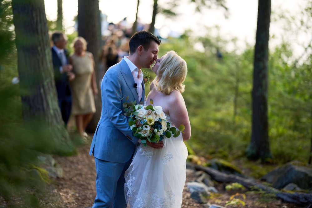 Newlyweds departing their lakeside ceremony along woodsy path pause for a happy kiss
