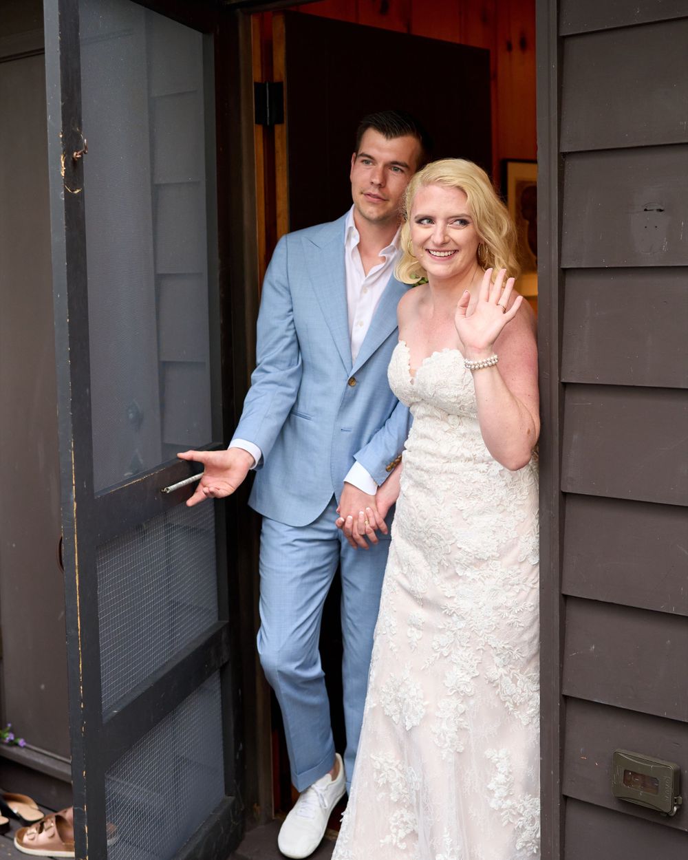 Newlyweds exit by lake house screen door to greet their guests with a happy wave