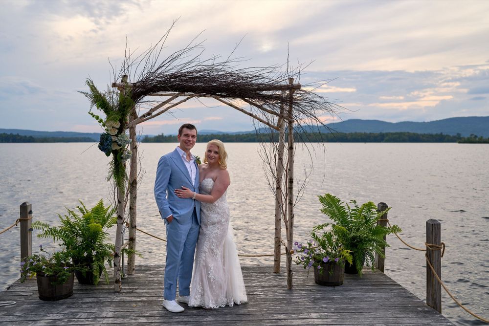 A cloud streaked sky frames newlyweds standing together at end of Squam Lake pier