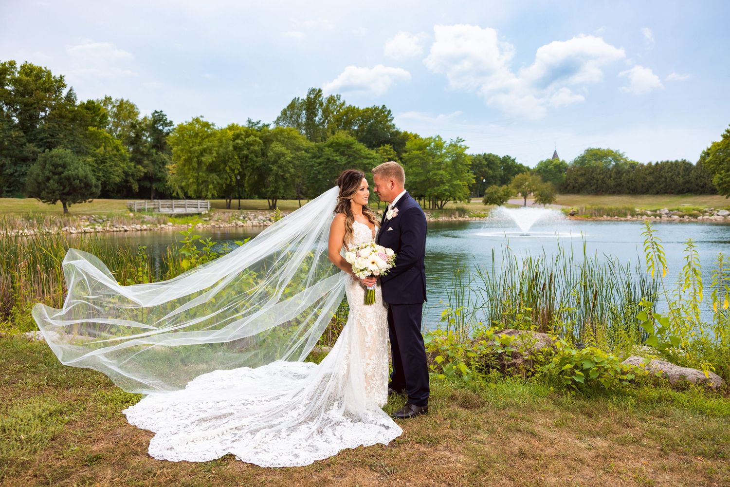Wedding couple embraces by lake with dramatic flowing veil in natural outdoor setting.