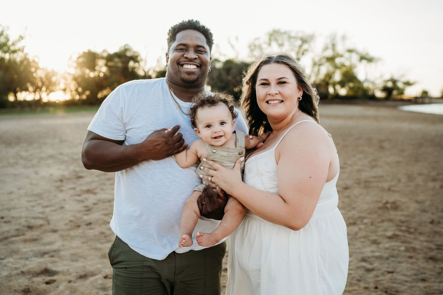 Family of three smiling together outdoors at sunset.