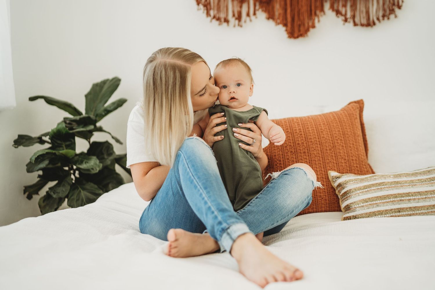 Parent and baby sharing tender moment on white bed with orange accents and indoor plants.