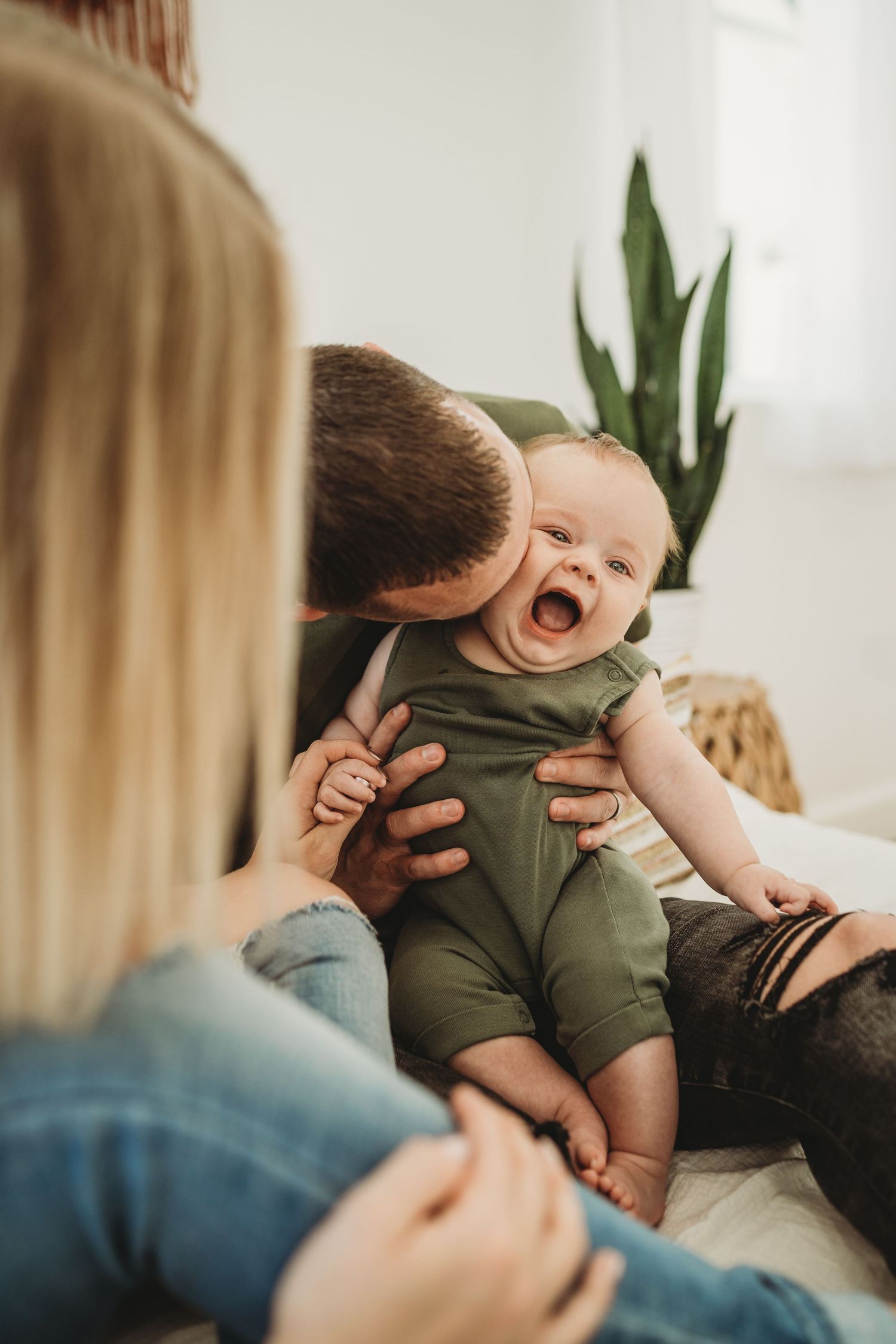 Laughing baby in olive green romper being held by parents on white bedding.