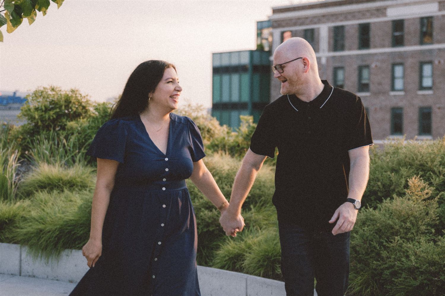 A couple holds hands while walking together on a rooftop garden with city buildings in background.