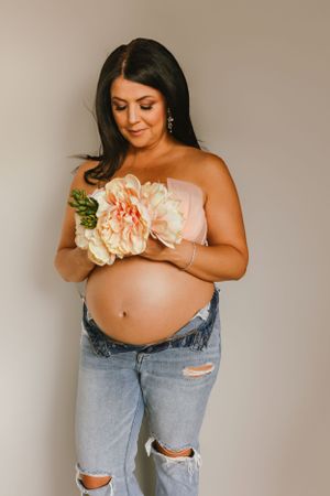 A maternity photo showing a person holding peach-colored flowers while wearing distressed jeans.