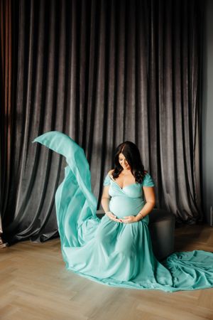 A maternity photo shoot features a flowing turquoise gown against a dark curtained backdrop.