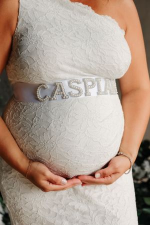 A close-up of a pregnant belly with a silver sash spelling 'CASPIE' over white lace dress.