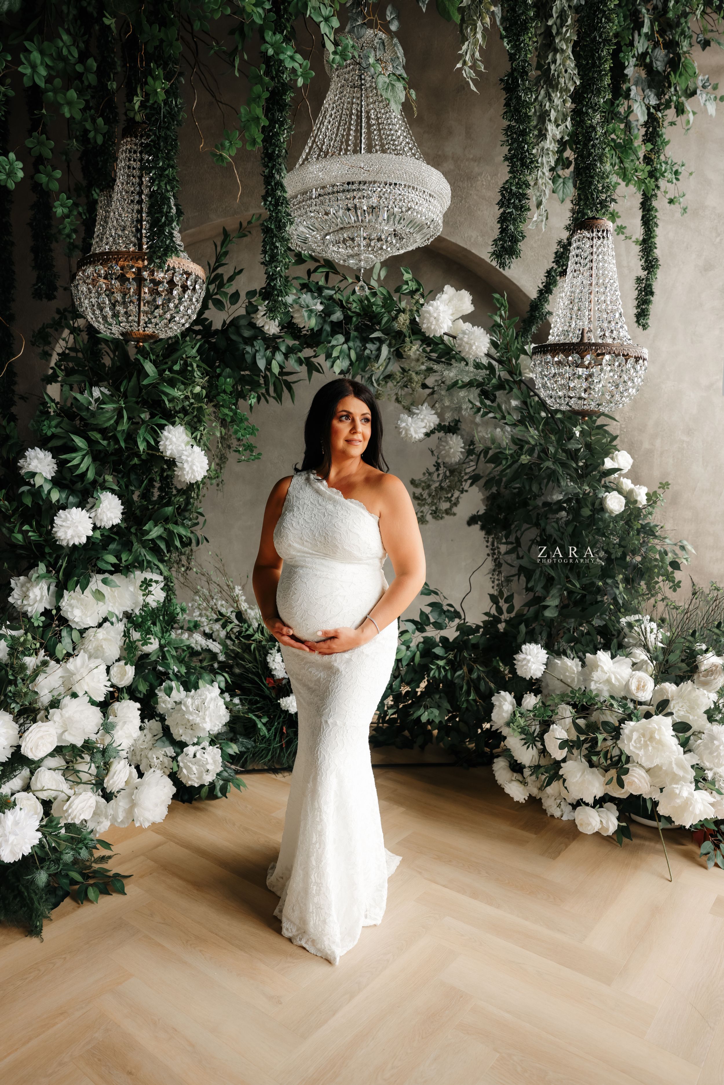 A maternity photo with crystal chandeliers and white floral arch creating an elegant backdrop.