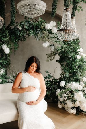 A maternity portrait under crystal chandeliers surrounded by white flowers and greenery.