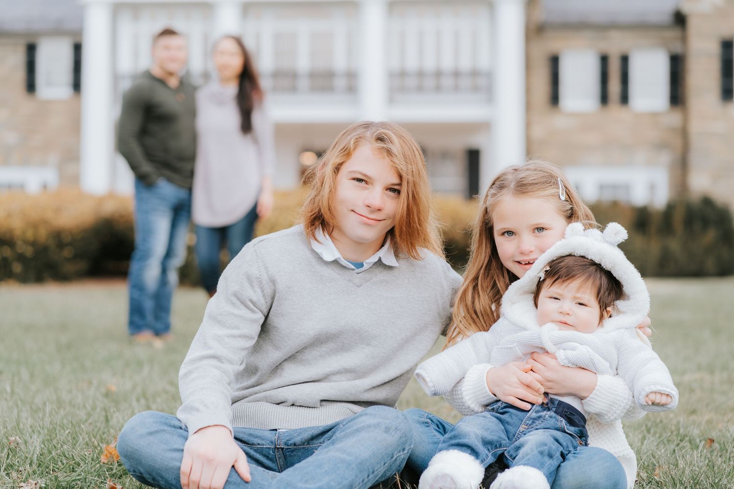 Family portrait with parents watching from background as children sit together in grass.