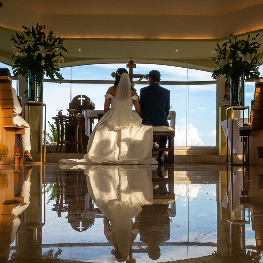Silhouette of couple seated at altar with large floral arrangements and waterfront view through windows.