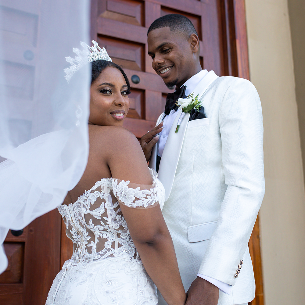 Newlyweds in elegant white attire share intimate moment beside wooden doors.