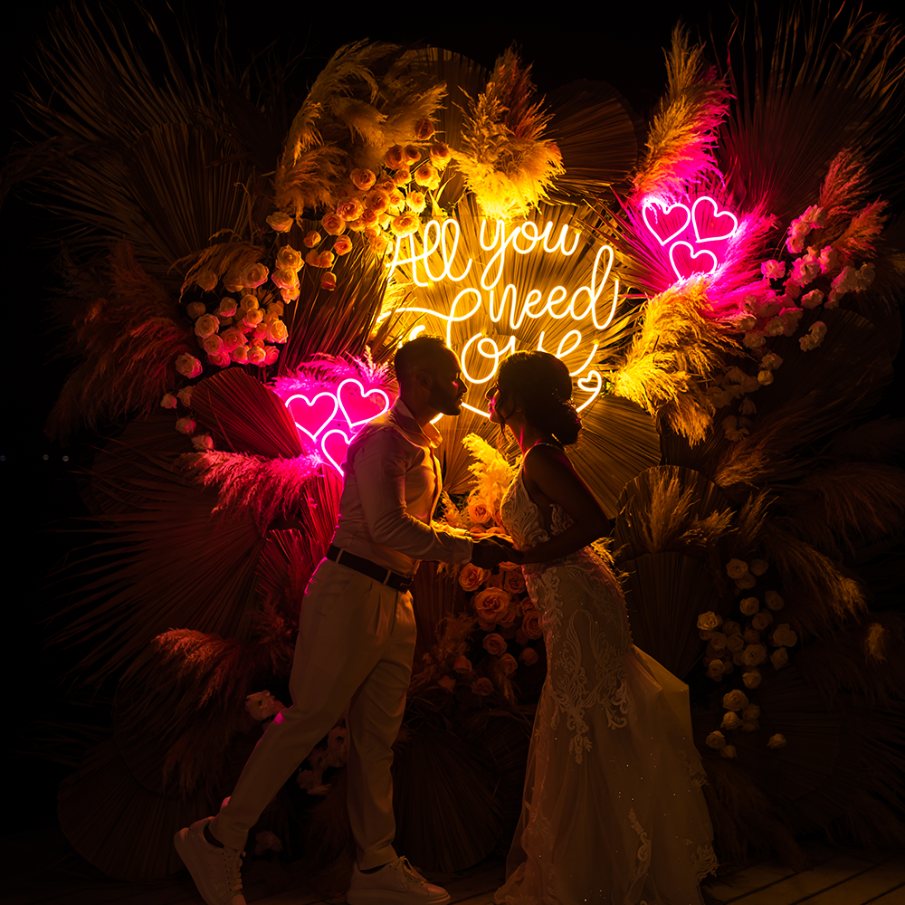 Couple embraces under glowing neon sign and floral arrangements at nighttime wedding.
