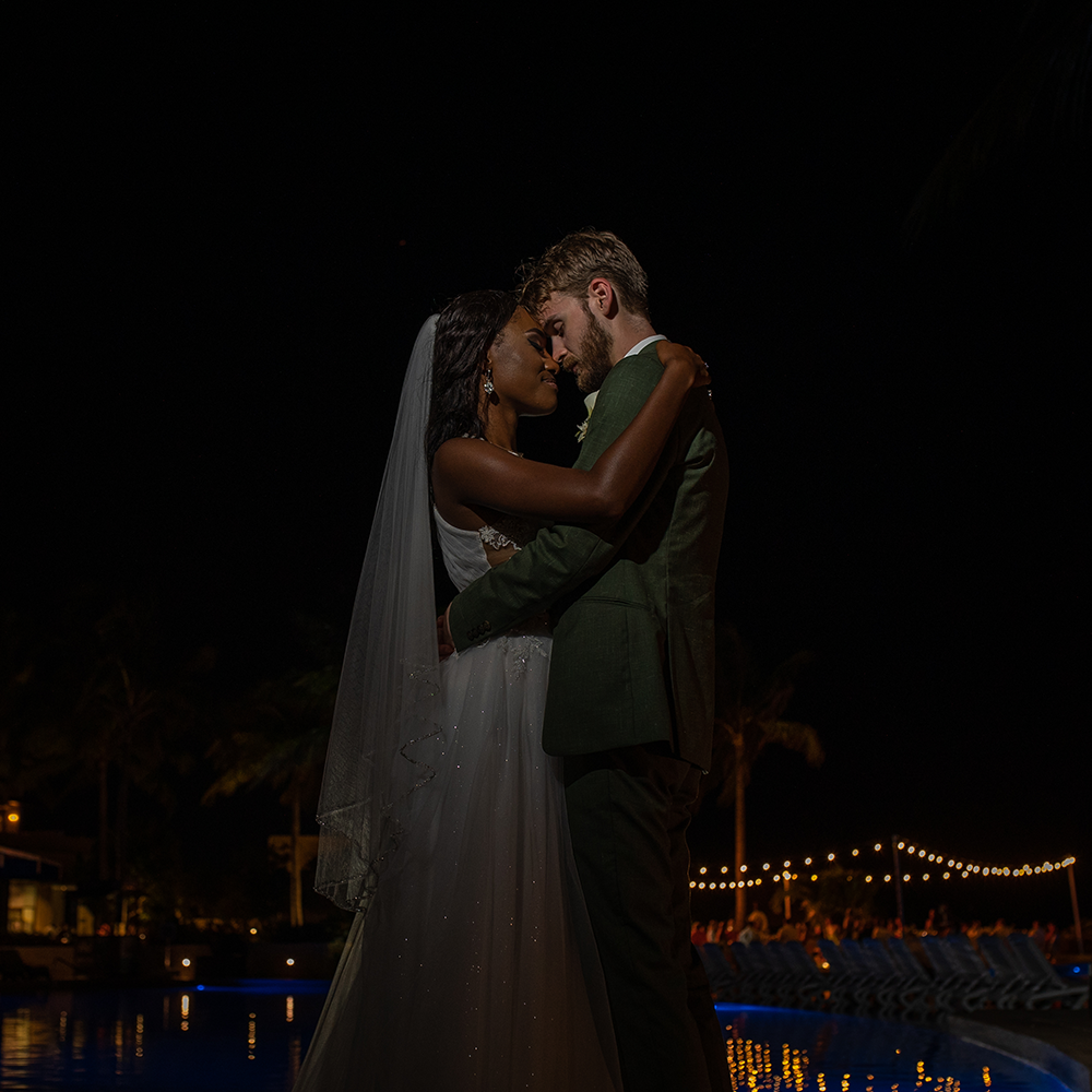 Romantic silhouette of couple embracing at night with string lights reflecting on water in background.