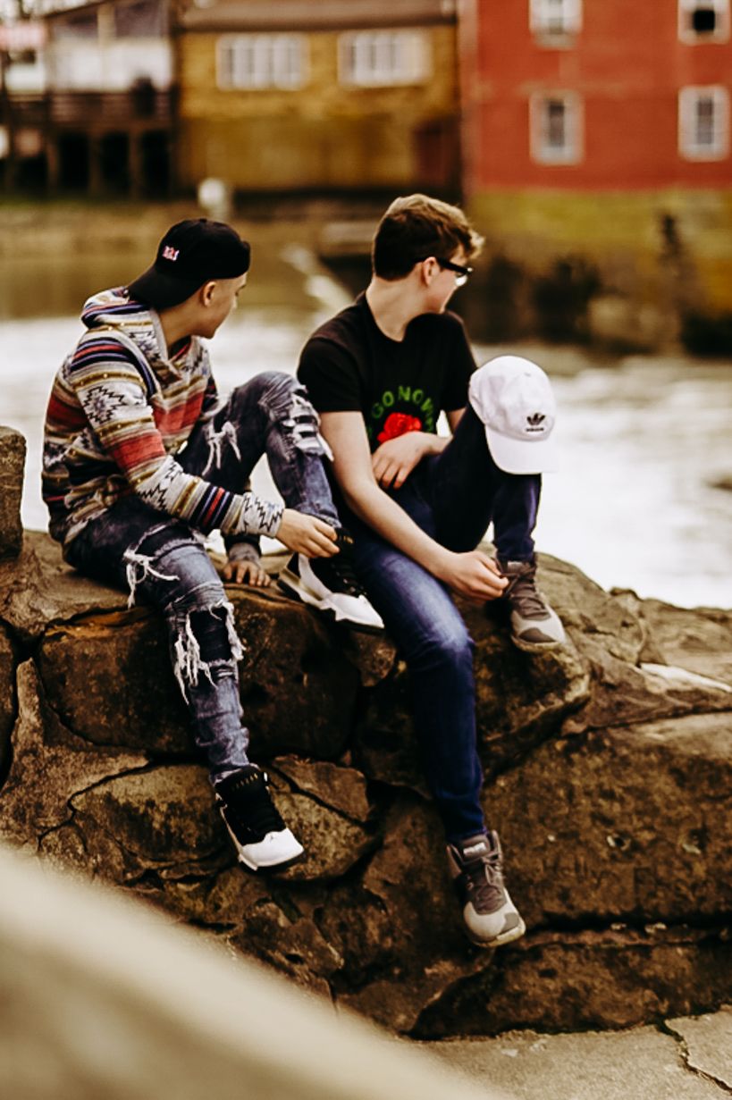 Two friends sitting on rocks by the water wearing casual streetwear and baseball caps.