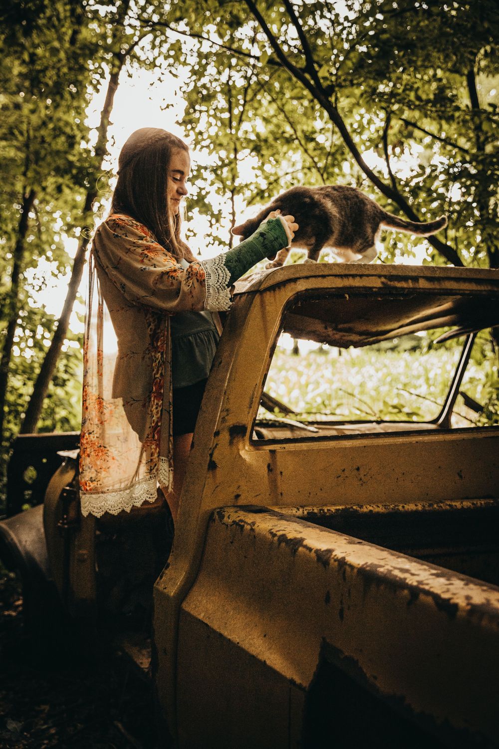 A woman in a flowy cardigan leans against an old rusty car in a wooded setting.