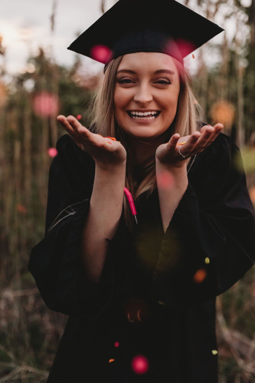 A graduate in cap and gown poses with outstretched hands while smiling outdoors.