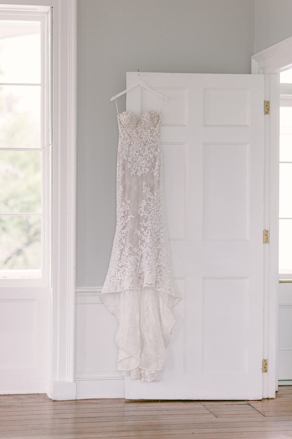 An elegant lace wedding dress hanging on display in a bright white room with hardwood floors.