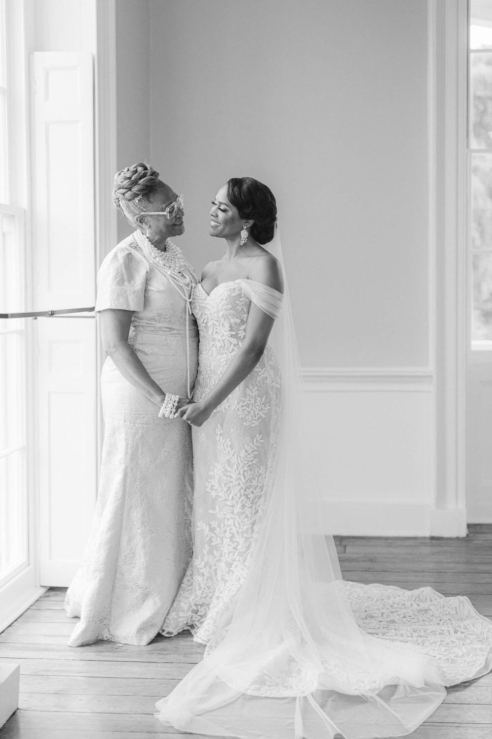 Two people in elegant white wedding dresses share an intimate moment by a bright window.