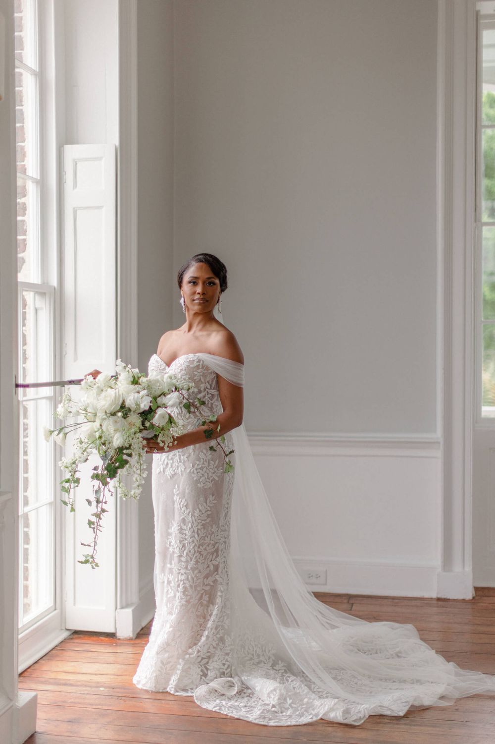 A person in an off-shoulder lace wedding gown holds a cascading white bouquet near a window.