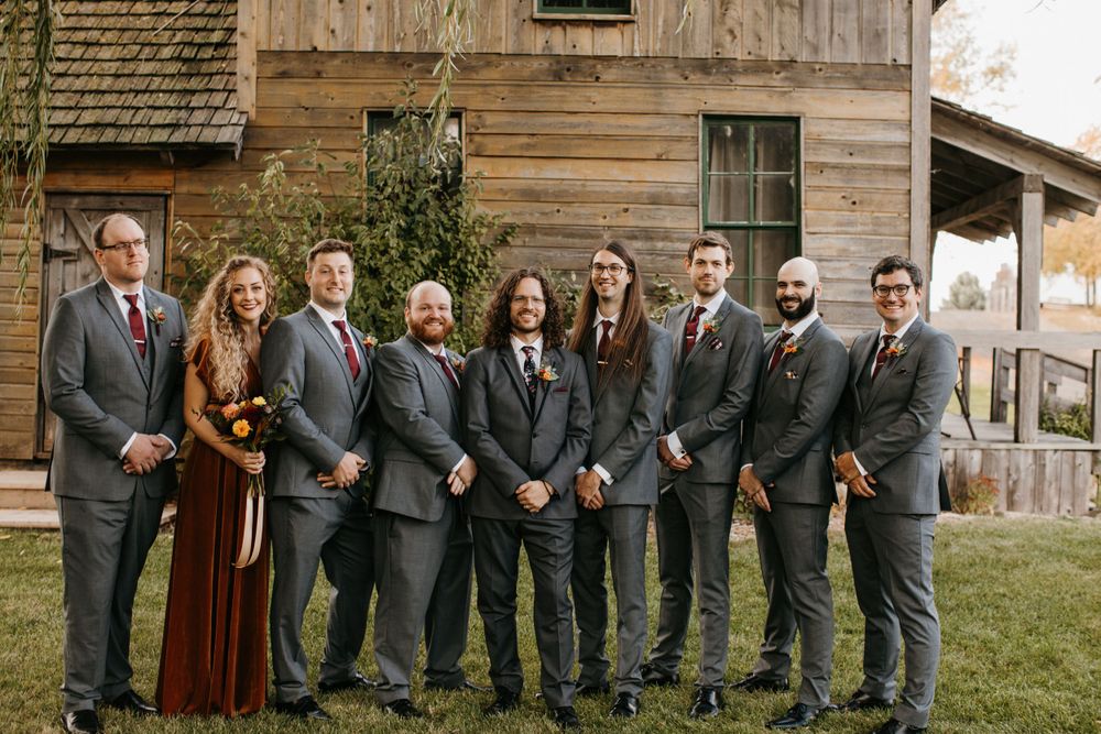 Groom and groomsmen in gray suits with burgundy ties pose before rustic wooden barn.