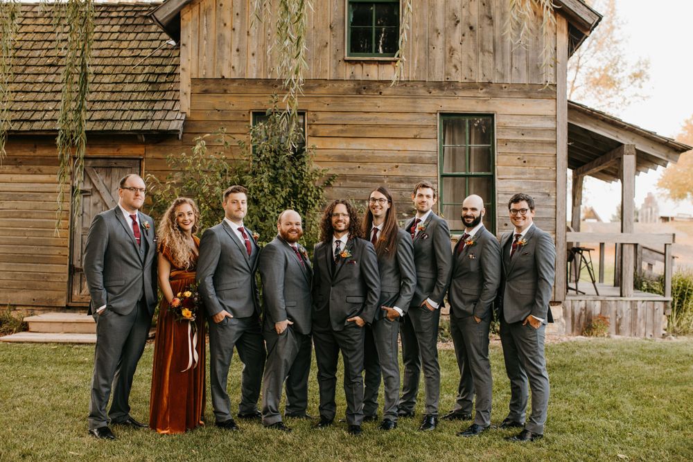 Groom with groomsmen in charcoal suits and maid of honor in burgundy dress at rustic barn.