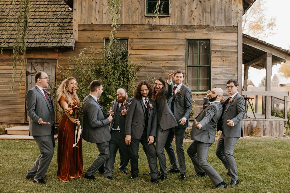 Wedding party laughing and posing casually in front of a rustic wooden barn in autumn.