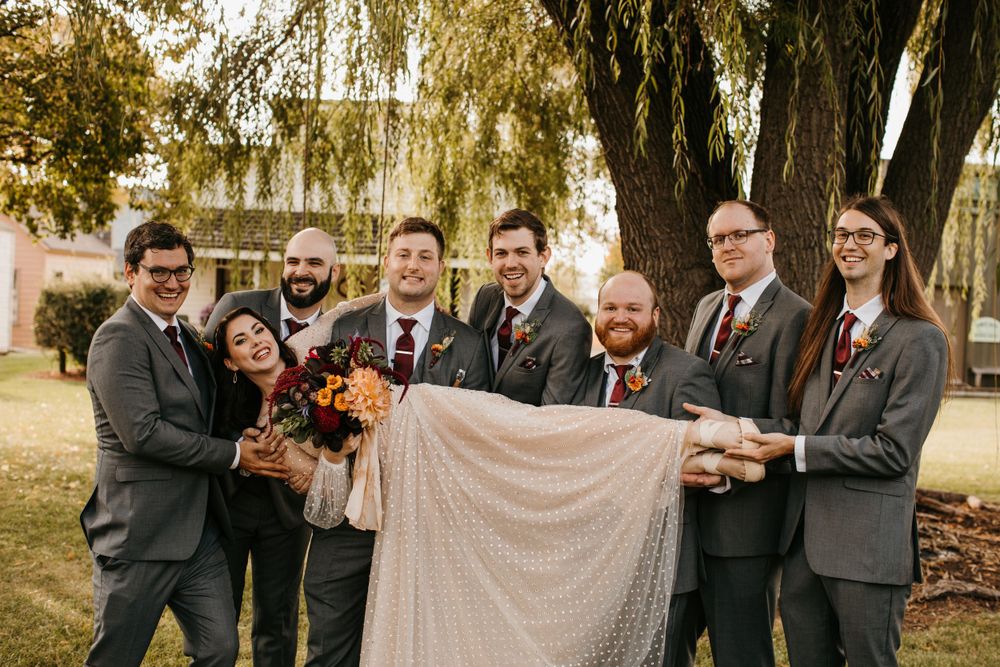 Bride and groom surrounded by wedding party holding bouquet under a large autumn tree.