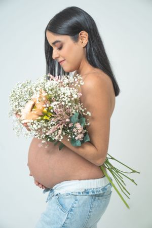 Side view of pregnant woman holding fresh bouquet against white studio background.